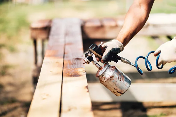 Uomo industriale che dipinge legno marrone con pistola a spruzzo e compressore — Foto Stock