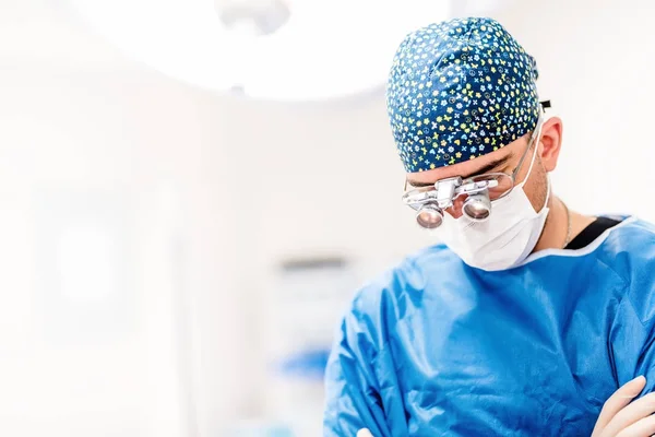 Porträtt av manliga kirurg, kosmetiska plastician tittar ner med kirurgisk lampor i bakgrunden. Läkare med luppar Royaltyfria Stockbilder