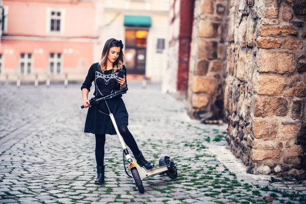 Hermosa joven mujer escribiendo mensaje de texto mientras monta scooter eléctrico en las calles urbanas — Foto de Stock