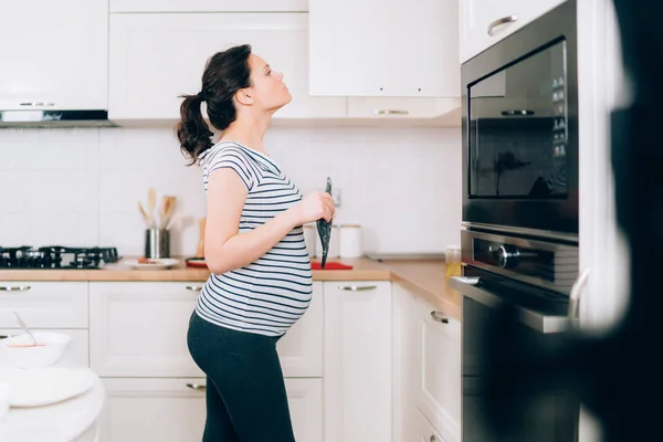 Mujer embarazada joven cocinando en su cocina de pie y mirando en los gabinetes cerca de la estufa — Foto de Stock