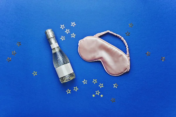 Bottle of champagne and mask for sleeping on blue background, pajama party concept — Stock Photo, Image