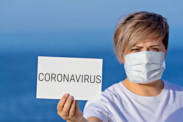 Woman in protective surgical mask holds blank with text coronavirus 2019