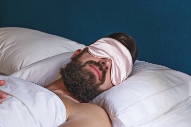 Göz maskeli beyaz sakallı adam yatakta uyuyor.