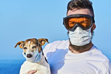 Koruyucu maskeli ve gözlüklü adam tıbbi maskeli sevimli Jack Russell köpeğini tutuyor. Evcil hayvan alerjisi ya da viral hastalıkların önlenmesi ve koronavirüs salgınının önlenmesi, kameraya bakmak, beyaz üzerinde izole edilmiş.