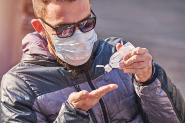 Maskeli bir adam parkta otururken ellerine antibakteriyel jel sürüyor. Coronavirus salgını sırasında kişisel önlemler.