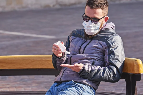 Maskeli bir adam parkta otururken ellerine antibakteriyel jel sürüyor. Coronavirus salgını sırasında kişisel önlemler.