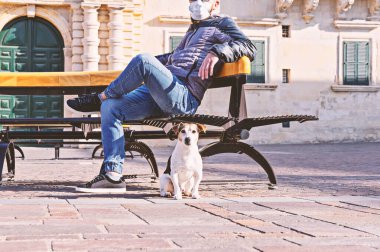 Parkta Coronavirus sırasında maskeli bir adam köpekle Becnh 'in üzerinde oturuyor. Covid-19 salgını sırasında kısa yürüyüşler. Karantina köpek kiralama konsepti