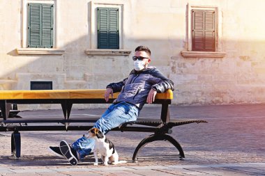 Parkta Coronavirus sırasında maskeli bir adam köpekle Becnh 'in üzerinde oturuyor. Covid-19 salgını sırasında kısa yürüyüşler. Karantina köpek kiralama konsepti