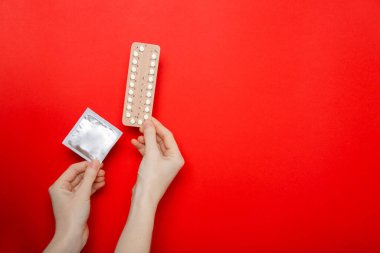 Kızın elinde doğum kontrol hapları ve prezervatifler var. Doğum kontrol hapı. Kırmızı arkaplan, metin için yer.