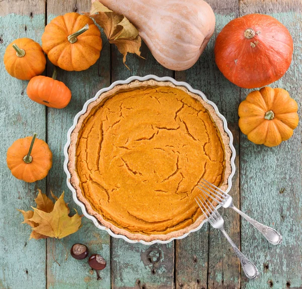 Homemade American pumpkin pie in white ceramic dish surrounded with small pumpkins, marple leaves and chestnuts on blue wooden background