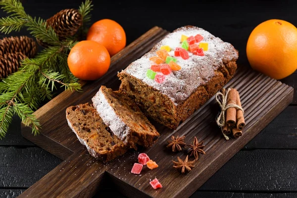 Tasty chocolate fruit cake decorated with candied fruits, clementines, spices and fir branch on carved dark oak board