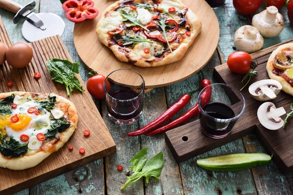 Homemade pizza party. Pizzas with eggs, spinach, mushrooms and chili served with raw vegetables and red wine on dark oak chopping boards over blue rustic background