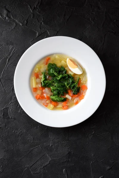 Cooking with local wild herbs. Tasty homemade soup with stringing nettle, vegetables and egg in white plate on black background top view copy space — Stock Image Cooking Local Wild Herbs Tasty Homemade Soup Stringing Nettle Vegetables Stock Image