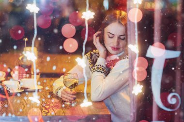 Genç ve güzel bir kadın kafede oturmuş, şarap içiyor. Sihirli kar yağışı etkisi. Noel, yeni yıl, sevgililer günü, kış tatili konsepti. Fotoğraf pencereden çekilmiş.