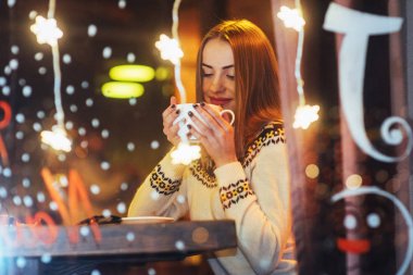 Kafede oturan, kahve içen genç ve güzel bir kadın. Sihirli kar yağışı etkisi. Noel, yeni yıl, sevgililer günü, kış tatili konsepti. Fotoğraf pencereden çekilmiş.
