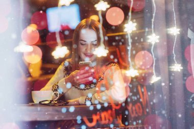 Kafede oturan, kahve içen genç ve güzel bir kadın. Sihirli kar yağışı etkisi. Noel, yeni yıl, sevgililer günü, kış tatili konsepti. Fotoğraf pencereden çekilmiş.