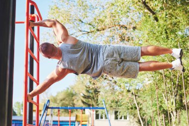 Güzel gövdesi olan kaslı bir adam bulanık park arka planında yatay parmaklıklar üzerinde egzersiz yapıyor. Delikanlı dışarıda barfiks çekiyor..