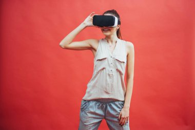 Mutlu gülümseyen güzel bir kızın portresi, kırmızı arka planda izole edilmiş sanal gerçeklik gözlüklerini kullanarak deneyim kazanmak.