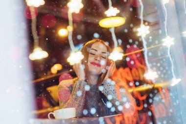 Kafede oturan, kahve içen genç ve güzel bir kadın. Müzik dinleyen bir model. Sihirli kar yağışı etkisi. Noel, mutlu yıllar, sevgililer günü, kış tatili konsepti. Fotoğraf pencereden çekilmiş.