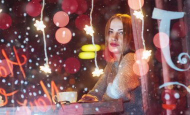 Kafede oturan, kahve içen genç ve güzel bir kadın. Sihirli kar yağışı etkisi. Noel, yeni yıl, sevgililer günü, kış tatili konsepti. Fotoğraf pencereden çekilmiş.