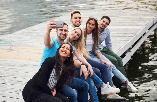 Grupa Młodych Ludzi Siedzących Skraju Molo Robiących Selfie Przyjaciele Cieszący — Zdjęcie stockowe