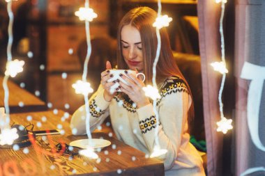 Kafede oturan, kahve içen genç ve güzel bir kadın. Sihirli kar yağışı etkisi. Noel, yeni yıl, sevgililer günü, kış tatili konsepti. Fotoğraf pencereden çekilmiş.