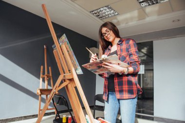 Güzel Güzel Kız ressamı sehpadaki tuvale resim yapıyor. Stüdyo siyah arka plan. Uzun saçlı..