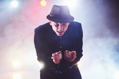 Sihirbaz iskambil kâğıdıyla numara yapıyor. Büyü ya da maharet, sirk, kumar. Karanlık odada sisli bir hokkabaz.