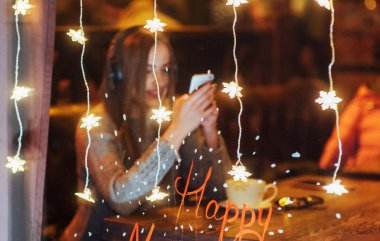 Kafede oturan, kahve içen genç ve güzel bir kadın. Müzik dinleyen bir model. Sihirli kar yağışı etkisi. Noel, mutlu yıllar, sevgililer günü, kış tatili konsepti. Fotoğraf pencereden çekilmiş.