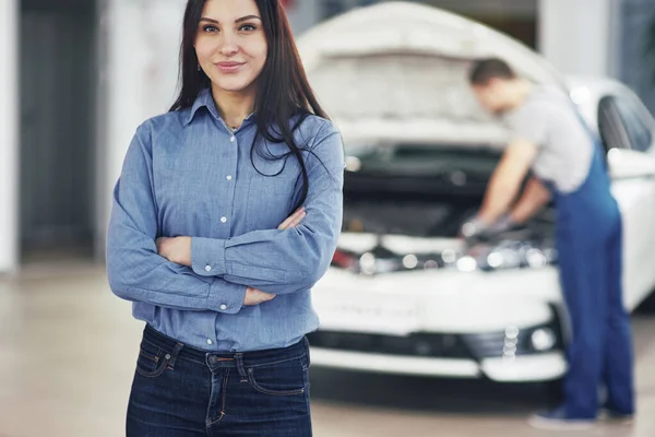 Woman at a car garage getting mechanical service. The mechanic works under the hood of the car.