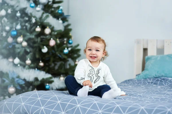 Very nice charming little girl blonde in white sitting on the bed and looking at the picture on the background of smiling Christmas trees in bright interior of the house.