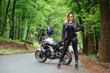 Deri giymiş çekici seksi bir kız dışarıda spor bir motosikletin yanında poz veriyor.