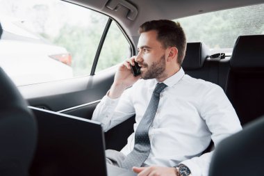 Genç bir iş adamı dizüstü bilgisayarda çalışıyor ve arabanın arkasında otururken telefonla konuşuyor. Hareket halinde çalışır, zamanını takdir eder..
