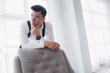 Klasik giysiler içinde genç yakışıklı bir adam ve beyaz pencerelere yaslanırken sandalyeye yaslanan gözlüklü..