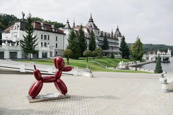 Luxury place. Monument of the dog made from balloons stands on the stand. Beautiful buildings, lake and forest at background.