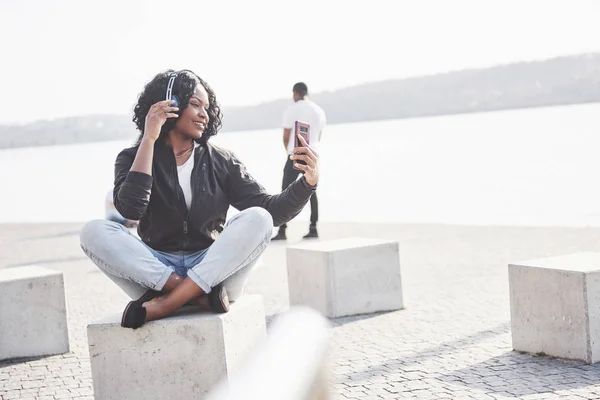 一个年轻漂亮的非洲裔美国女孩坐在海滩或湖上 用耳机听音乐的画像 — 图库照片