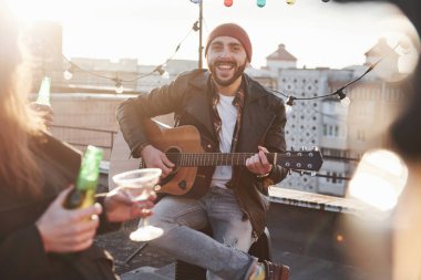 Akustik gitarlı mutlu adam kameraya gülümsüyor ve arkadaşlarıyla çatıda oturuyor..
