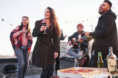 Şaka yapıyordum. Çatıda gülümseyen ve içen neşeli gençler. Masada pizza ve alkol var. Gitarist.