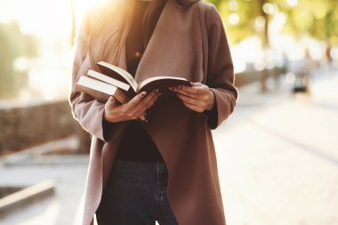 Genç, yüzü olmayan esmer kız elinde küçük bir yığın kitap tutarken ve kahverengi bir kitap okurken sonbahar parkında arka planda ağaçlarla fotokopi çektirirken..