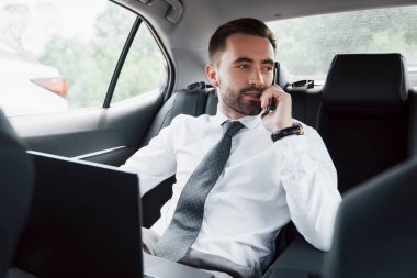Genç bir iş adamı dizüstü bilgisayarda çalışıyor ve arabanın arkasında otururken telefonla konuşuyor. Hareket halinde çalışır, zamanını takdir eder..