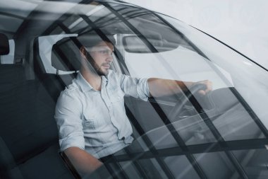 Genç bir adam yeni alınmış bir arabada oturuyor ve ellerini dümene koyuyor.