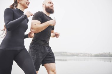 Couple jogging and running outdoors in park near the water. Young bearded man and woman exercising together in morning.