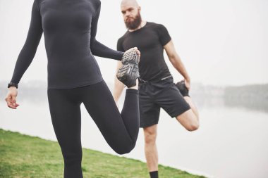 Fitness couple stretching outdoors in park near the water. Young bearded man and woman exercising together in morning.