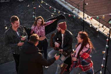Çekici esmer kız gülümseyip resme poz veriyor. İçki içen, gitar çalan ve dekoratif renkli ampullerle çatıda eğlenen genç bir arkadaşın en iyi görüntüsü..