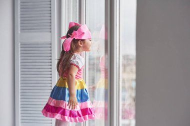 Çocukça bir merak. Renkli giyinmiş genç bir kızın fotoğrafı pencerenin yanında duruyor ve dışarı bakıyor..