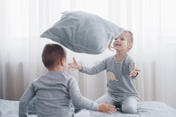 Happy kids playing in white bedroom. Little boy and girl, brother and sister play on the bed wearing pajamas. Nursery interior for children. Nightwear and bedding for baby and toddler. Family at home