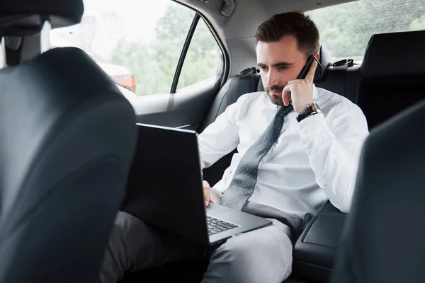 Młody Biznesmen Pracujący Laptopie Rozmawiający Przez Telefon Siedząc Tyłu Samochodu — Zdjęcie stockowe