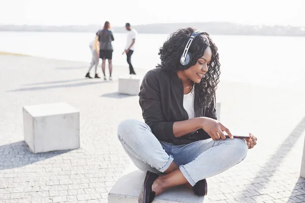 一个年轻漂亮的非洲裔美国女孩坐在海滩或湖上 用耳机听音乐的画像 — 图库照片