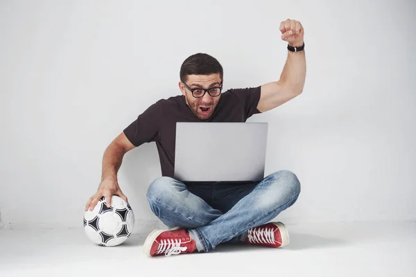 兴奋的足球迷 有着白色背景的足球 他坐在地板上 在笔记本电脑上观看比赛的结果和比赛的最佳时刻 — 图库照片