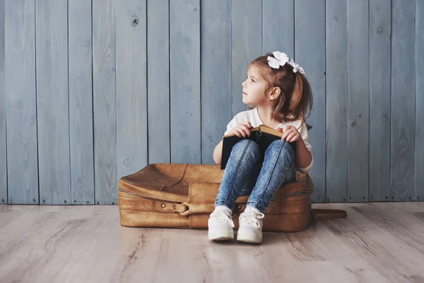 Ready to big travel. Happy little girl reading interesting book carrying a big briefcase and smiling. Travel, freedom and imagination concept.